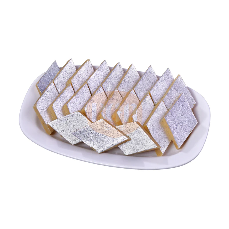 A white plate filled with diamond-cut pieces of Kaju Katli, dusted with edible silver leaf, showcasing their elegant shape and luxurious appearance